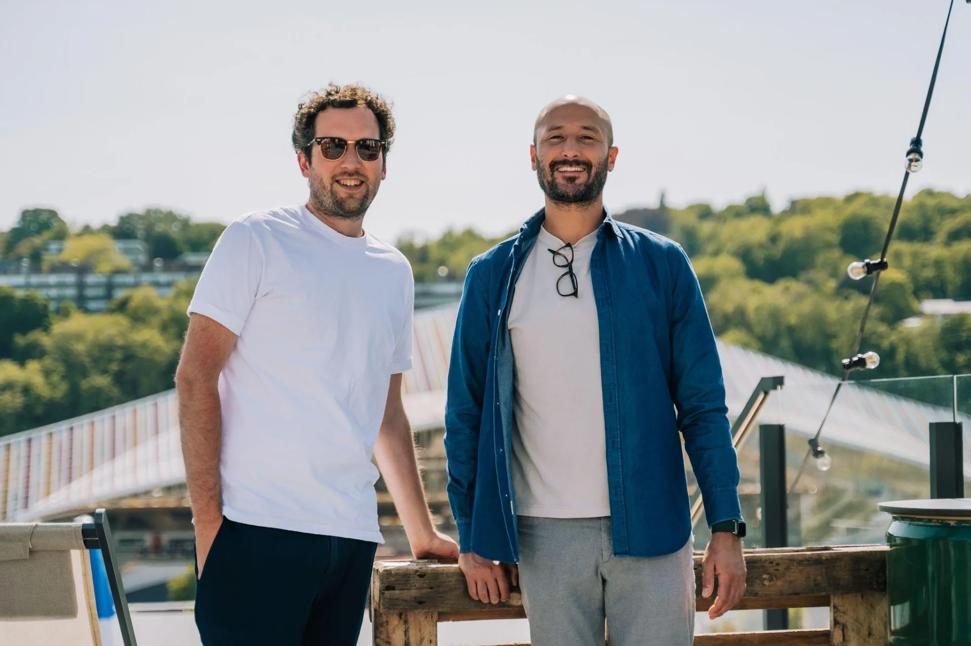 glooh's two co-founders on a rooftop terrace in the sun, trees and a bridge behind them