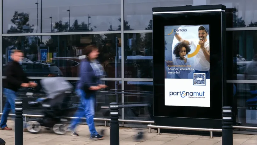 A Partenamut campaign running on a glooh screen outside a supermarket entrance, shoppers walking past
