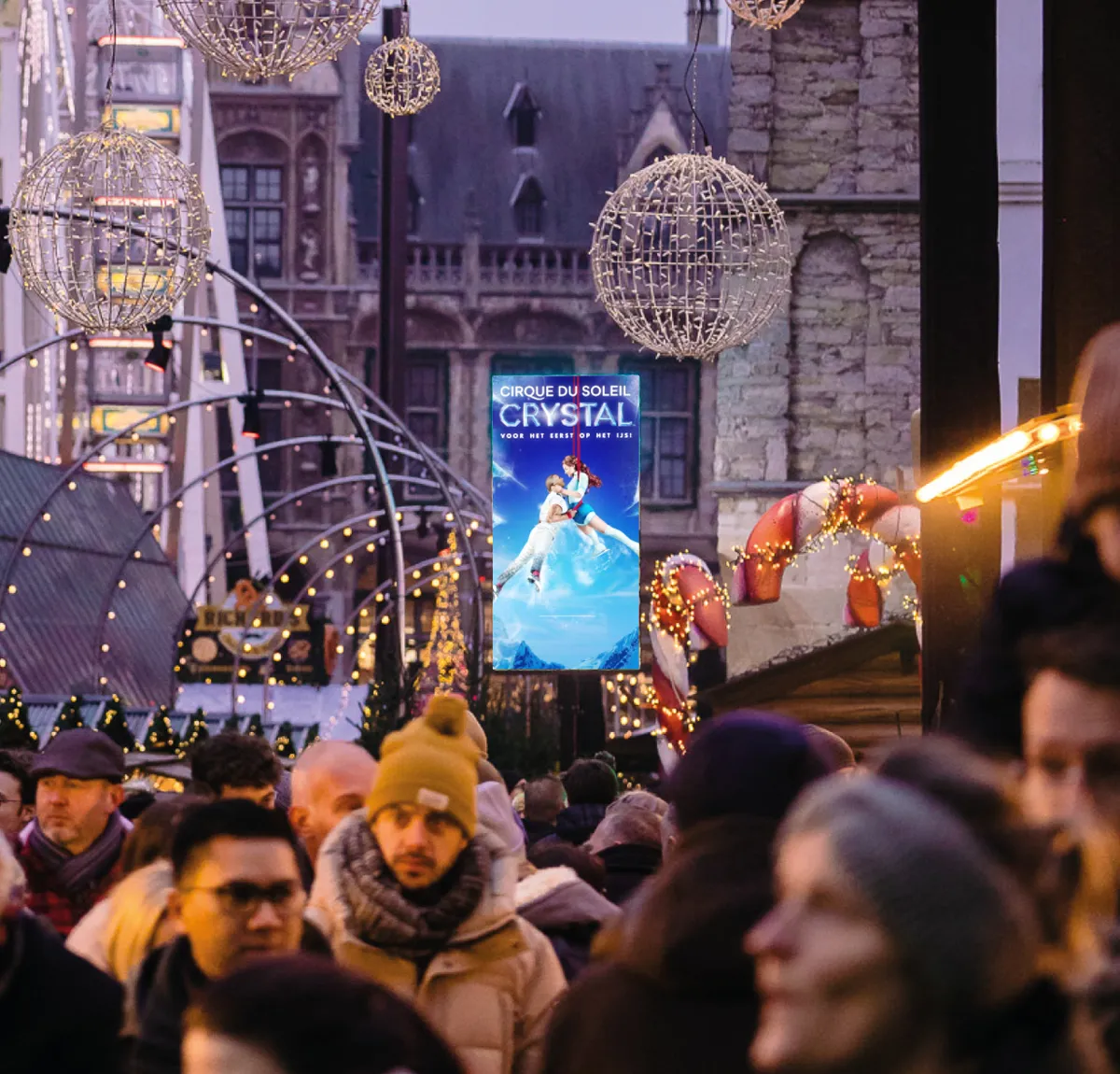 Digital screen showing an advert above the crowd in a Christmas market, chalets and light decorations behind
