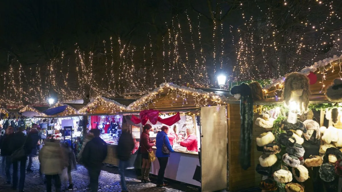 Christmas market chalets under strings of lights in the trees at night, visitors at a stall