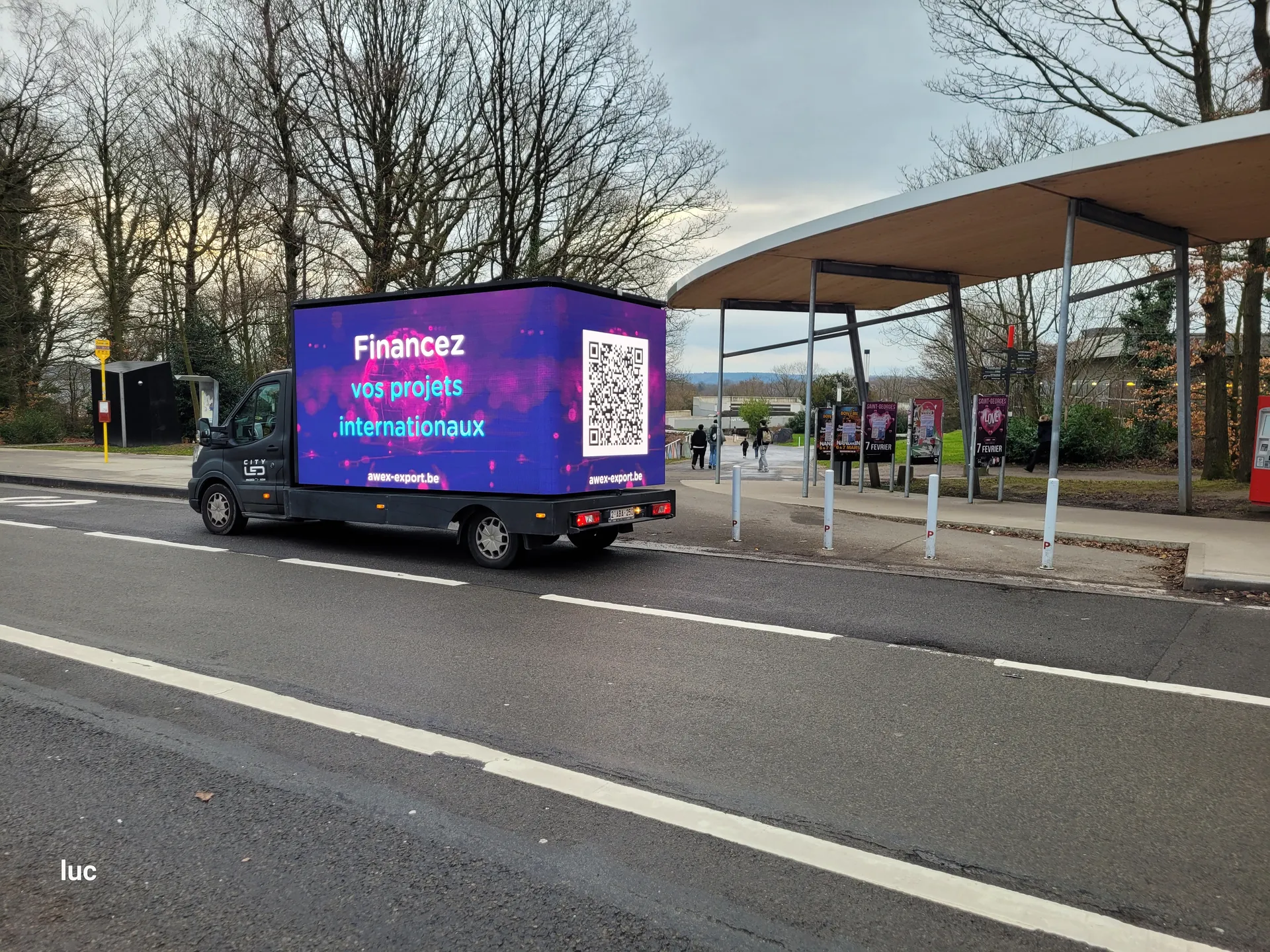 A glooh mobile LED truck carrying the AWEX export-incentive campaign, parked at a bus stop on the road into a Walloon business park